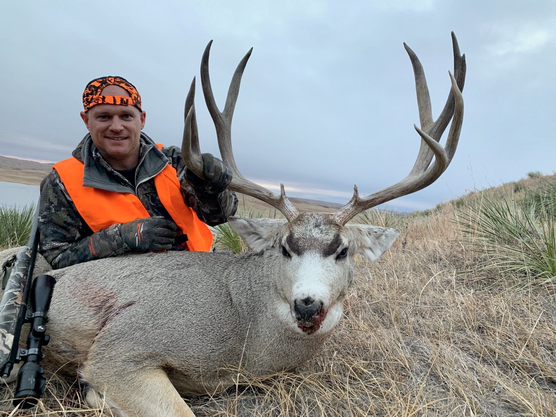 Hunter smiles, poses with large buck, both in grassy field. Hunter wears orange safety vest, buck has large antlers.
