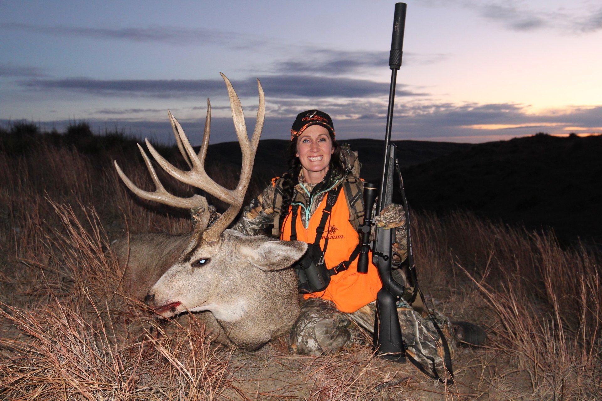 Woman in camo with a large buck she hunted, rifle leaned beside her, outdoors at dusk.