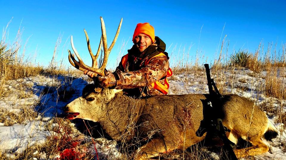 Hunter smiles, kneeling beside a large buck with large antlers, snow on ground, gun nearby, orange cap.