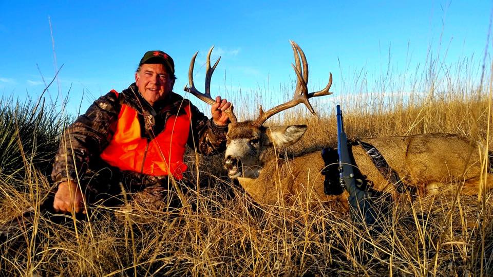 Hunter in orange vest kneels next to a large buck he shot; gun lies nearby in tall, dry grass.