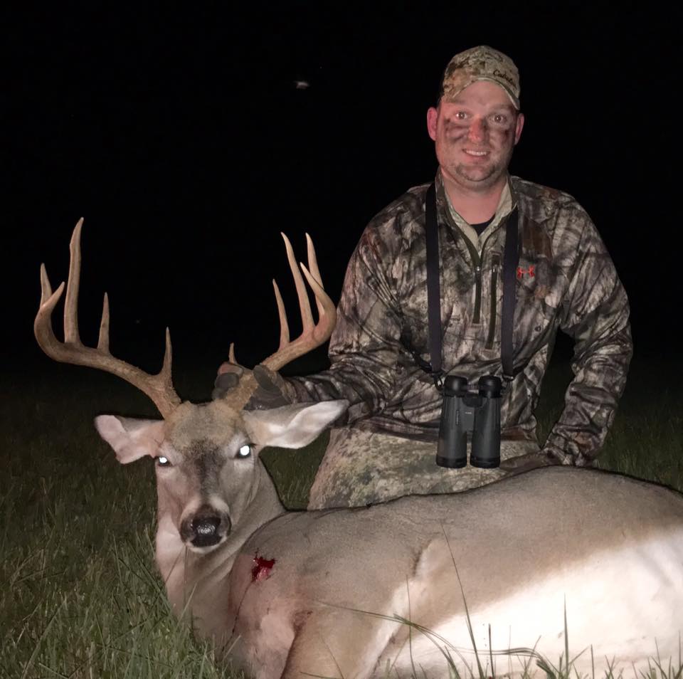 Man in camo next to a harvested buck. Night setting, deer has large antlers and a visible wound.
