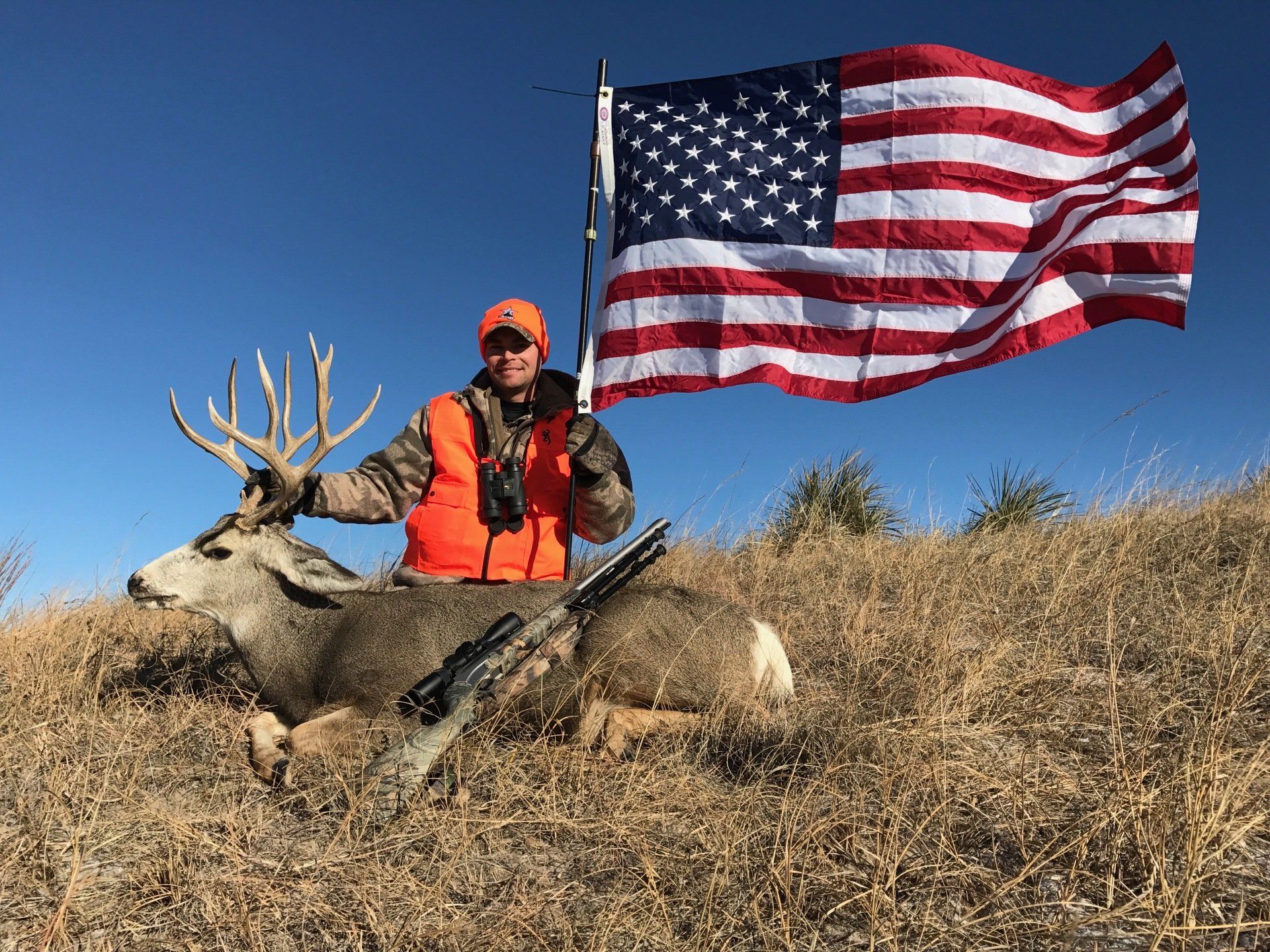 Hunter poses with a large deer, American flag in background, wearing orange vest, outdoors.