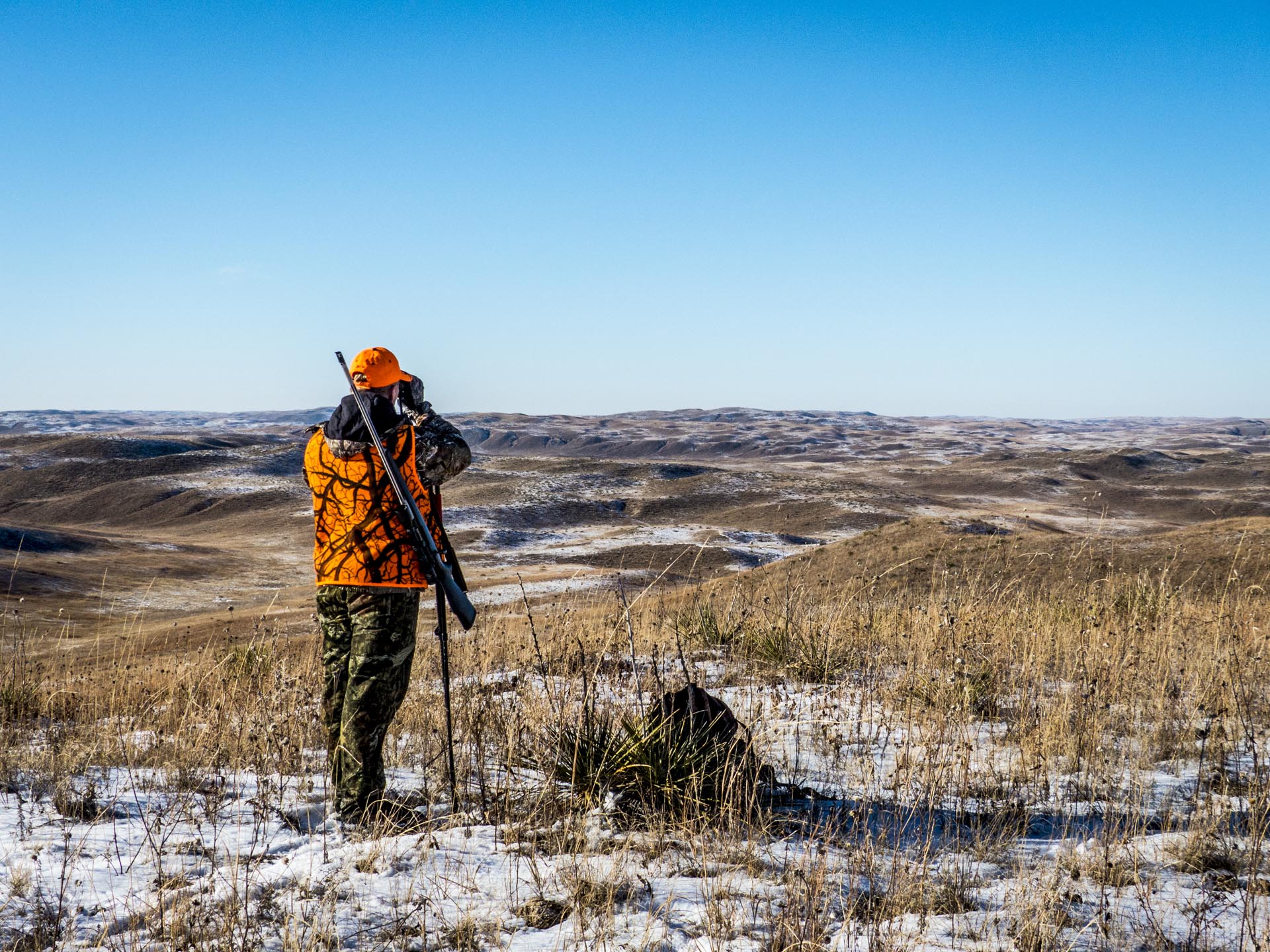 Hunter in orange vest and camo pants scans a snowy, hilly landscape with a rifle on a bipod.