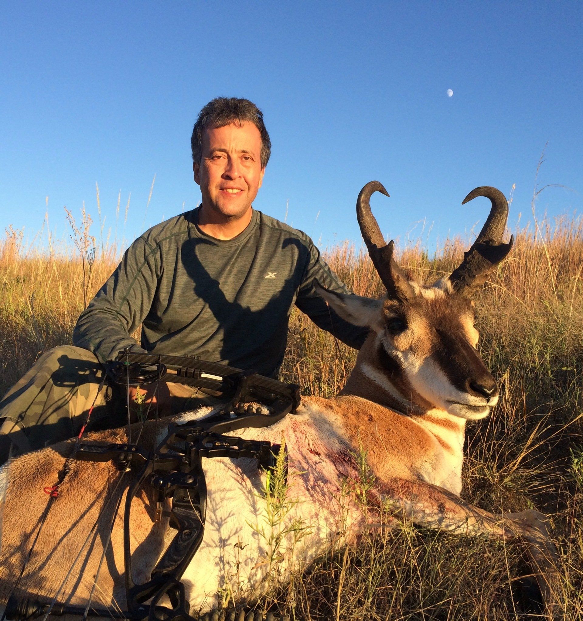 Man smiles next to a pronghorn antelope he hunted. Outdoor setting, clear sky. Bow is visible.