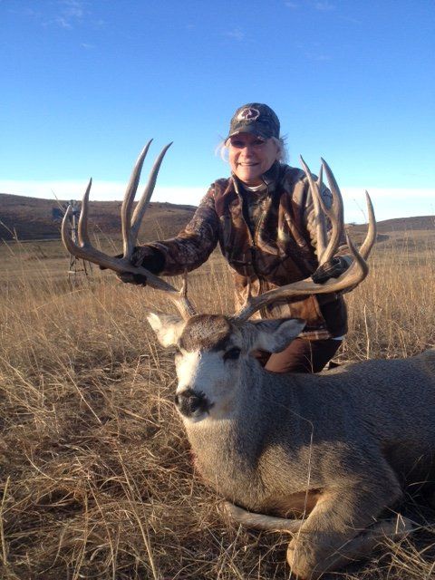 Woman in camouflage kneeling next to a large buck with impressive antlers in a field.