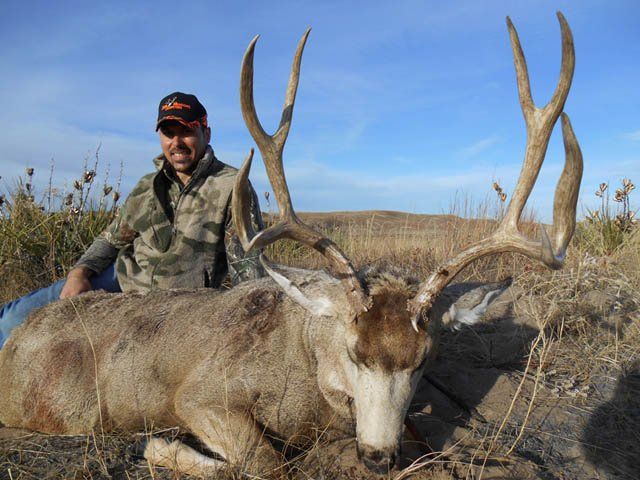 Hunter kneels next to a large dead buck with impressive antlers, outdoors under a blue sky.