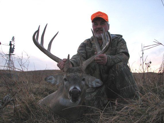 Hunter kneeling with a large deer buck, holding its antlers. Outdoors, overcast sky, camouflage, orange hat.