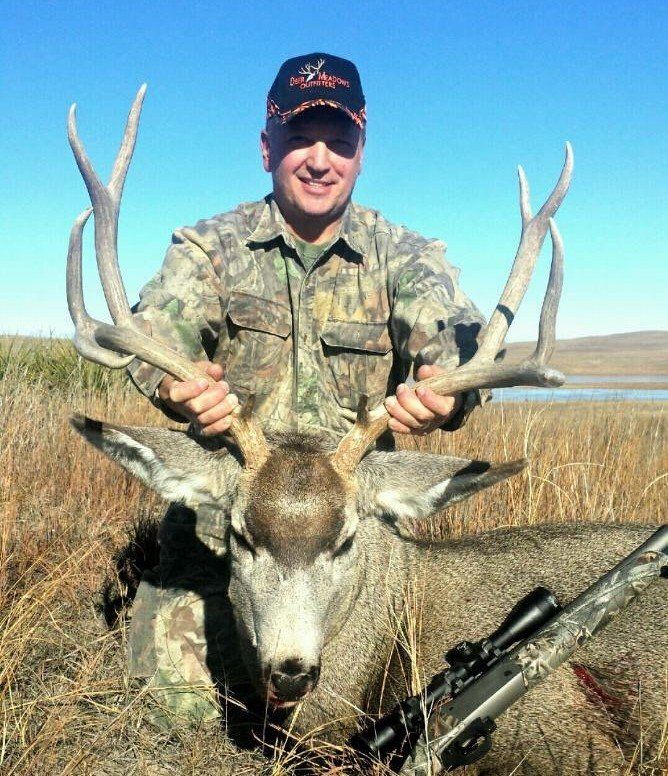 Man in camouflage holds deer antlers, smiling, with rifle in field.