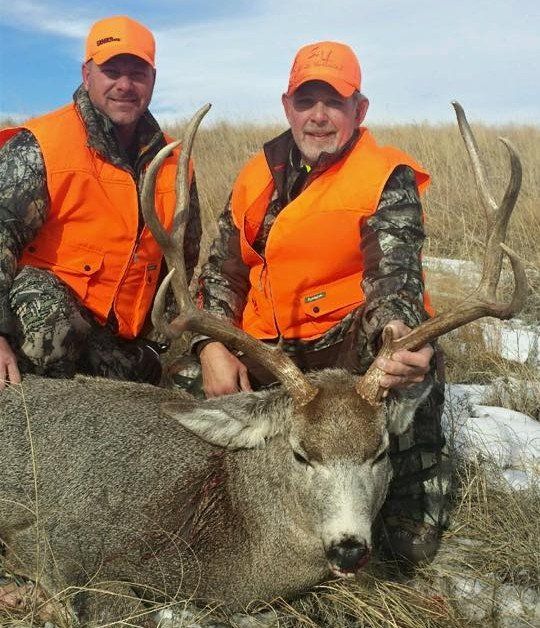 Two men in orange vests pose with a harvested deer in a grassy field.