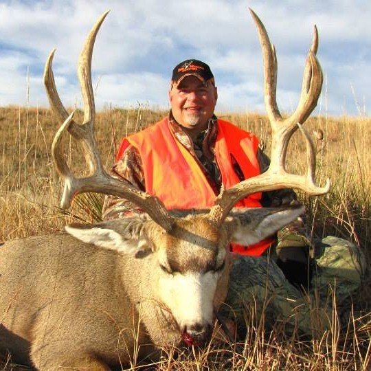 Hunter in orange vest kneels next to large buck with impressive antlers in a grassy field.