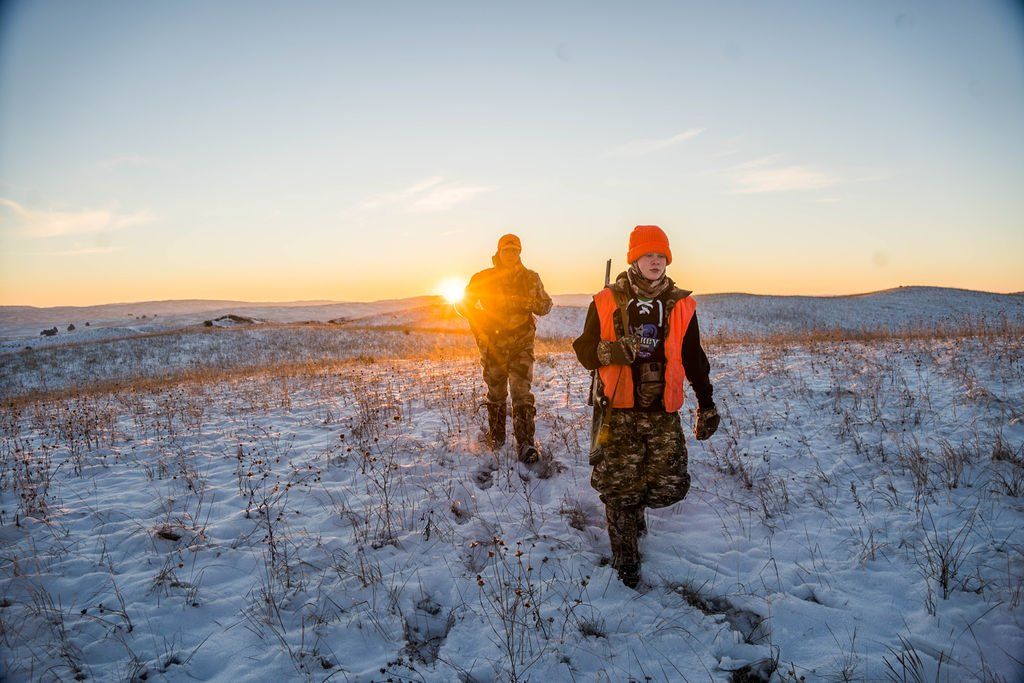 Two hunters walk across snow-covered field at sunset. One wears orange safety vest and camo, the other, a jacket.