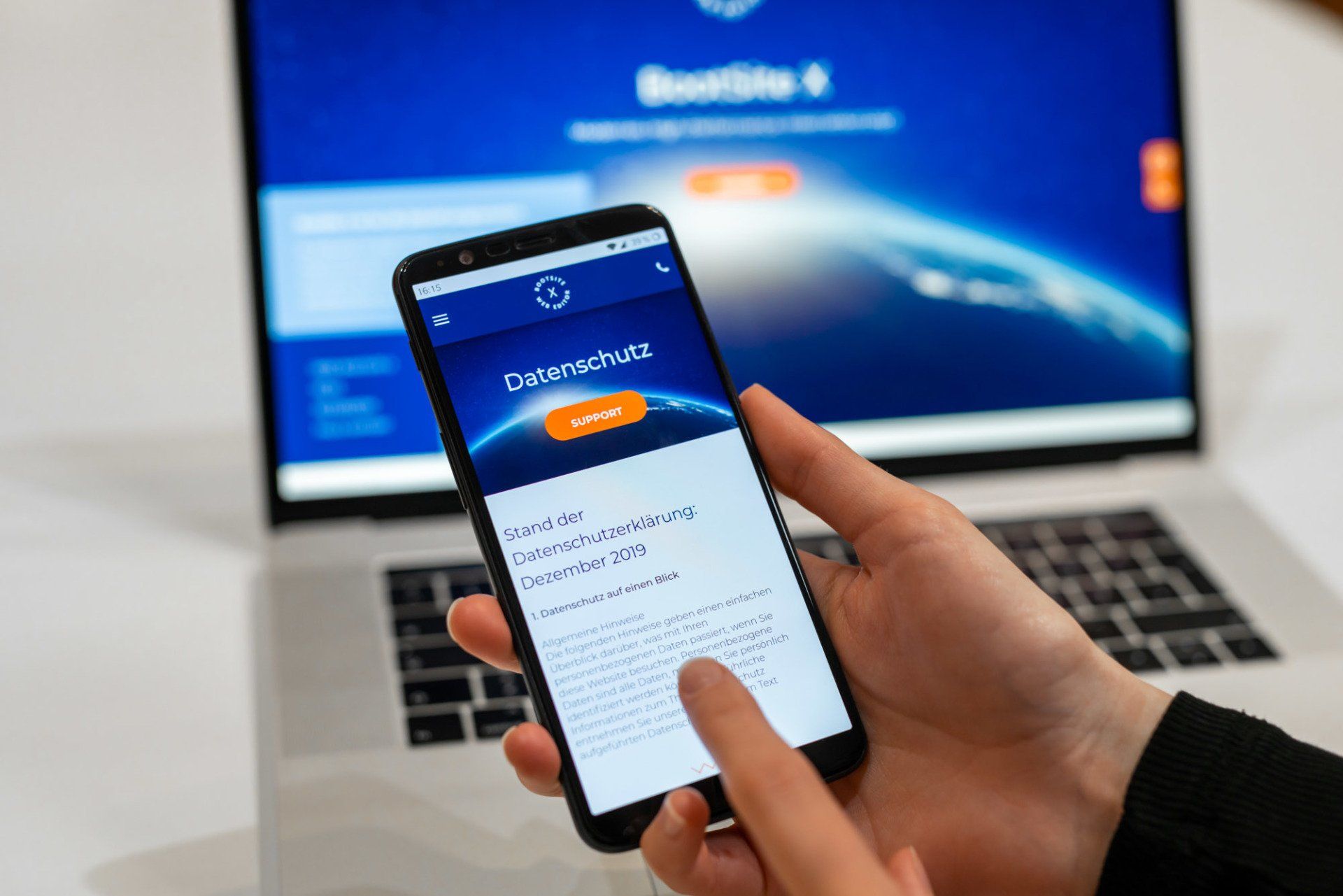 Eine Hand hält ein Smartphone mit Anmeldebildschirm vor einem Laptop, auf dem eine blaue Webseite geöffnet ist.