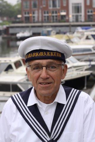 A person wearing a white sailor hat and traditional navy-collared uniform, smiling in front of a marina.