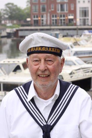 An individual in a white sailor uniform and cap smiles in front of a marina with docked boats.