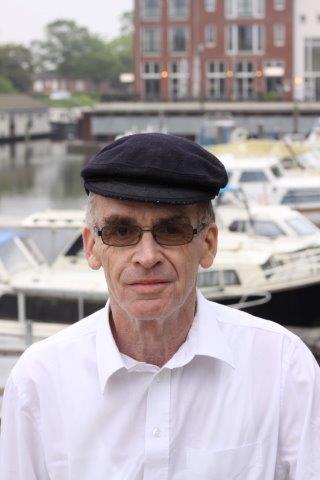 A person wearing a dark flat cap and glasses stands outdoors with a marina and brick buildings in the background.