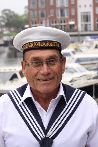 A smiling person in a white sailor uniform and cap with gold lettering, standing in front of a marina with boats.