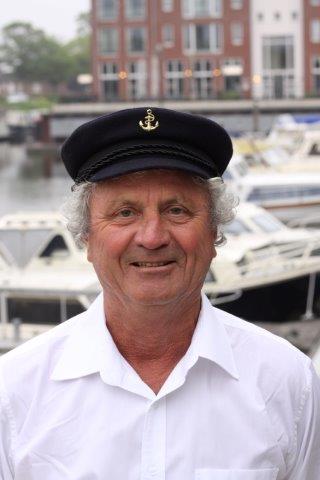 A smiling person wearing a white shirt and a navy captain's hat, positioned in front of a marina with boats and buildings.