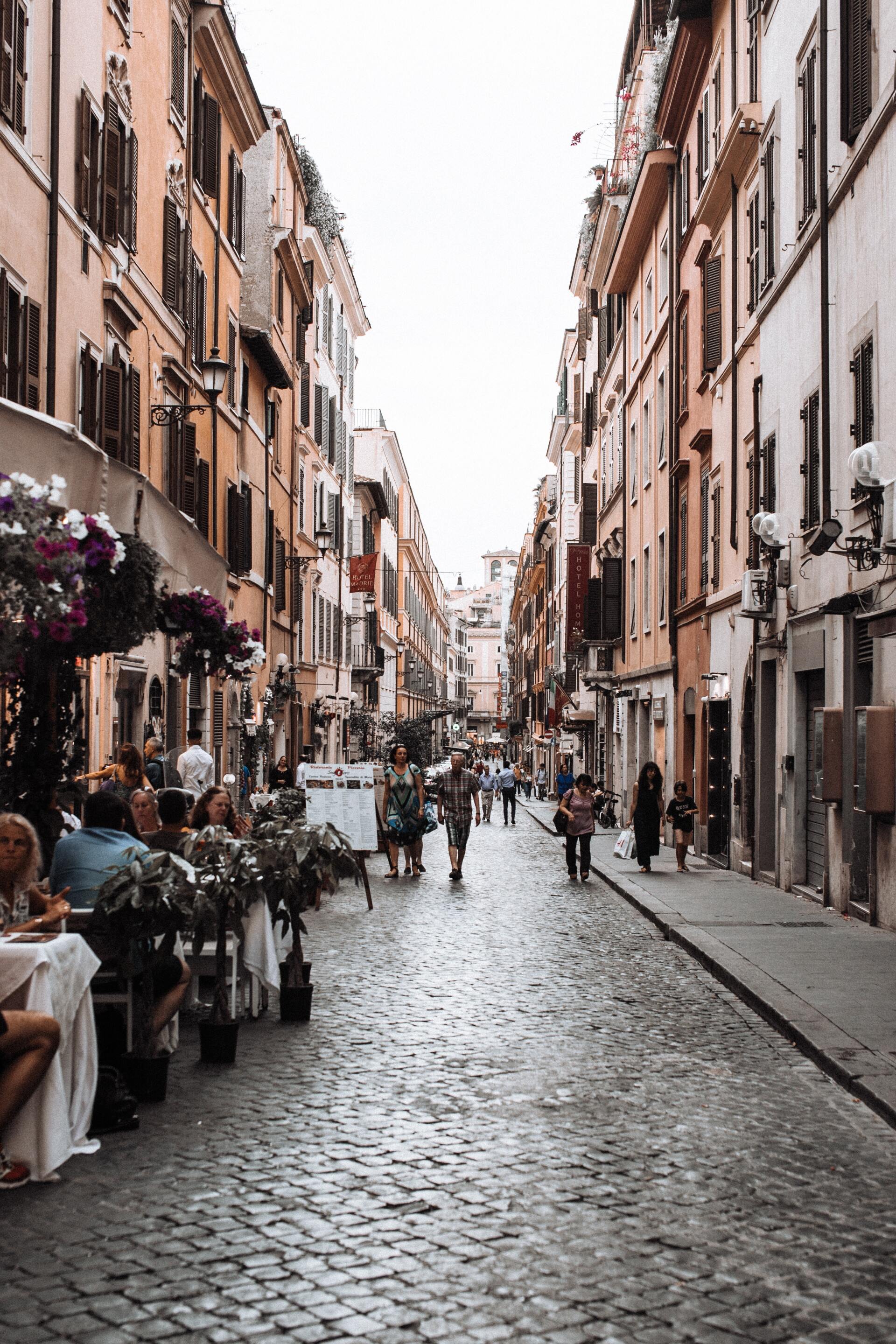 Cobblestone street lined with buildings, cafe tables, and people in Rome.