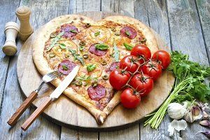 Hartvormige pizza met toppings op een houten plank met tomaten, peterselie, knoflook, zout- en pepervaatjes en bestek.