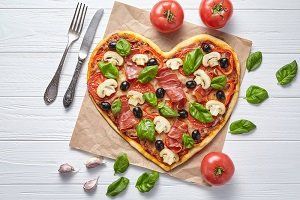 Hartvormige pizza met tomaten, basilicum, olijven en champignons op bakpapier; ernaast een vork en een mes.