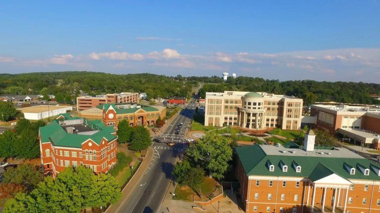 Aerial view of a tree-lined campus with red-brick and white buildings along a central road under a blue sky.