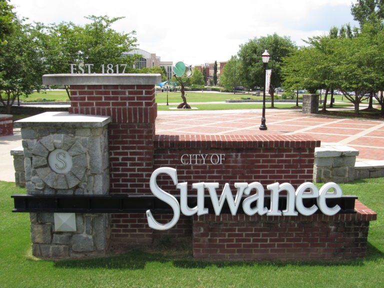 Suwanee city sign with brick and stone base in a park, trees and walkway in the background