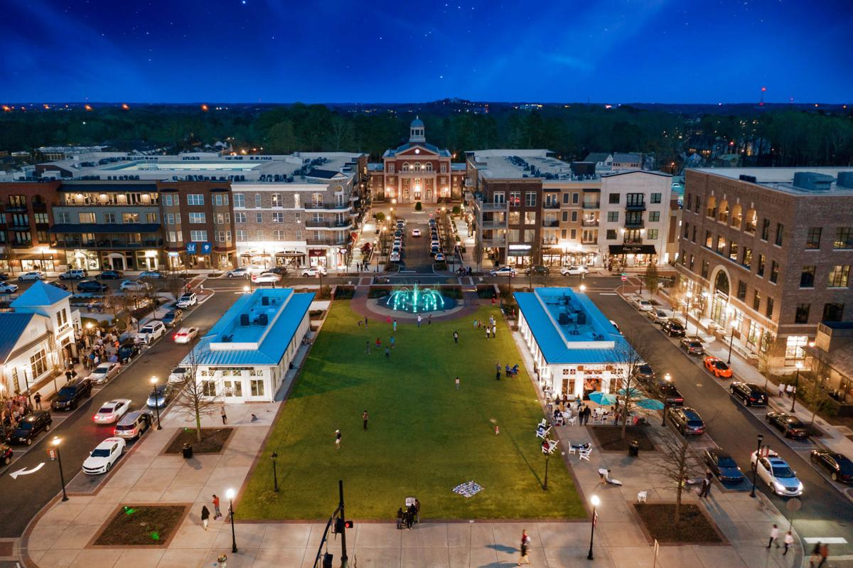 Aerial view of a lit city square with a green lawn, surrounding buildings, and a central monument at dusk