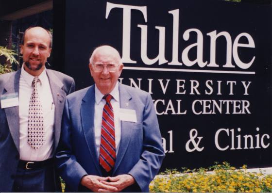 Two men in suits stand in front of a Tulane University Medical Center sign.