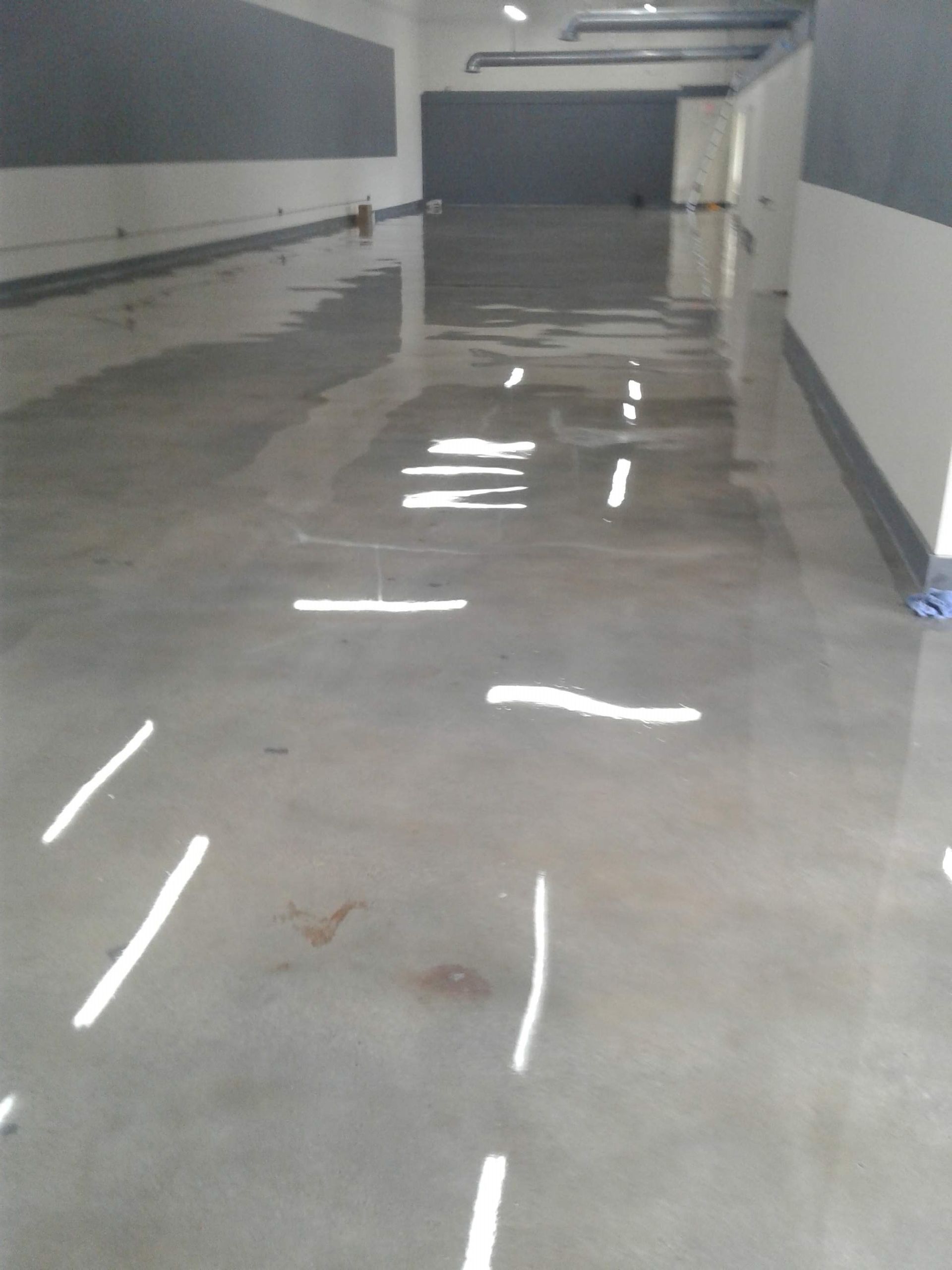 Empty indoor hallway with glossy wet floor reflecting lights, gray walls, and a dark doorway at the end