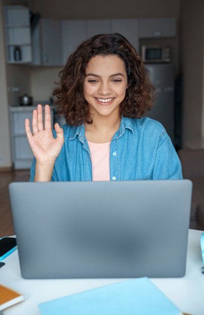 A high school student is waving at the camera while sitting in front of a laptop computer at a virtual college fair.