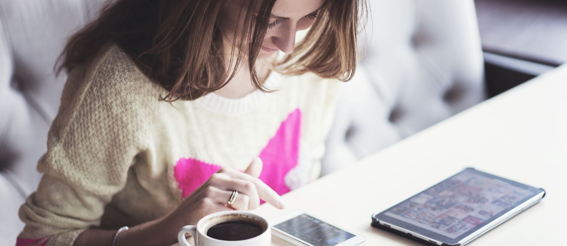 Eine Frau sitzt mit einer Tasse Kaffee und einem Tablet an einem Tisch.