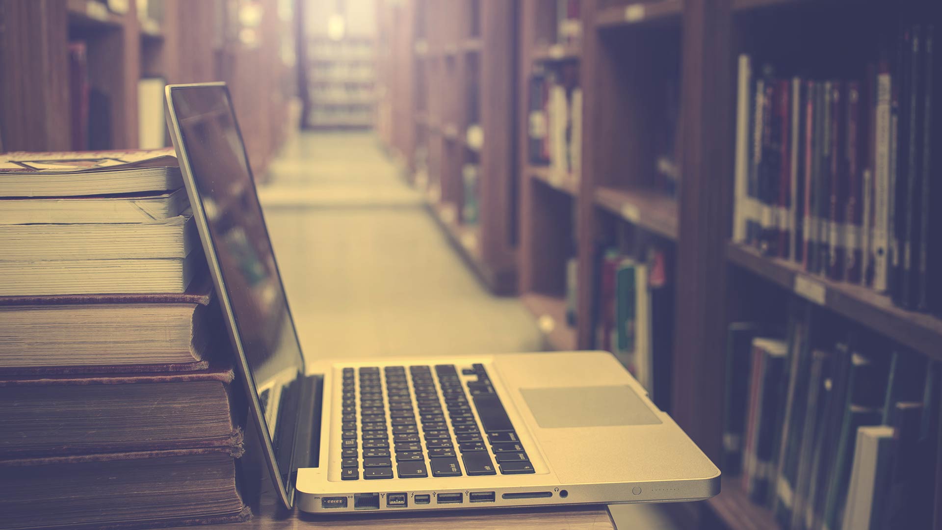 Um laptop está sobre uma mesa em uma biblioteca ao lado de uma pilha de livros.