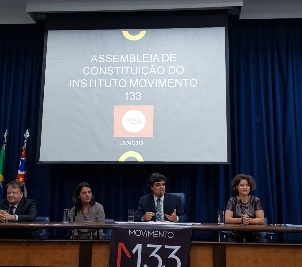 Um grupo de pessoas sentadas a uma mesa em frente a uma tela que diz assembleia de constituição do instituto movimento 133