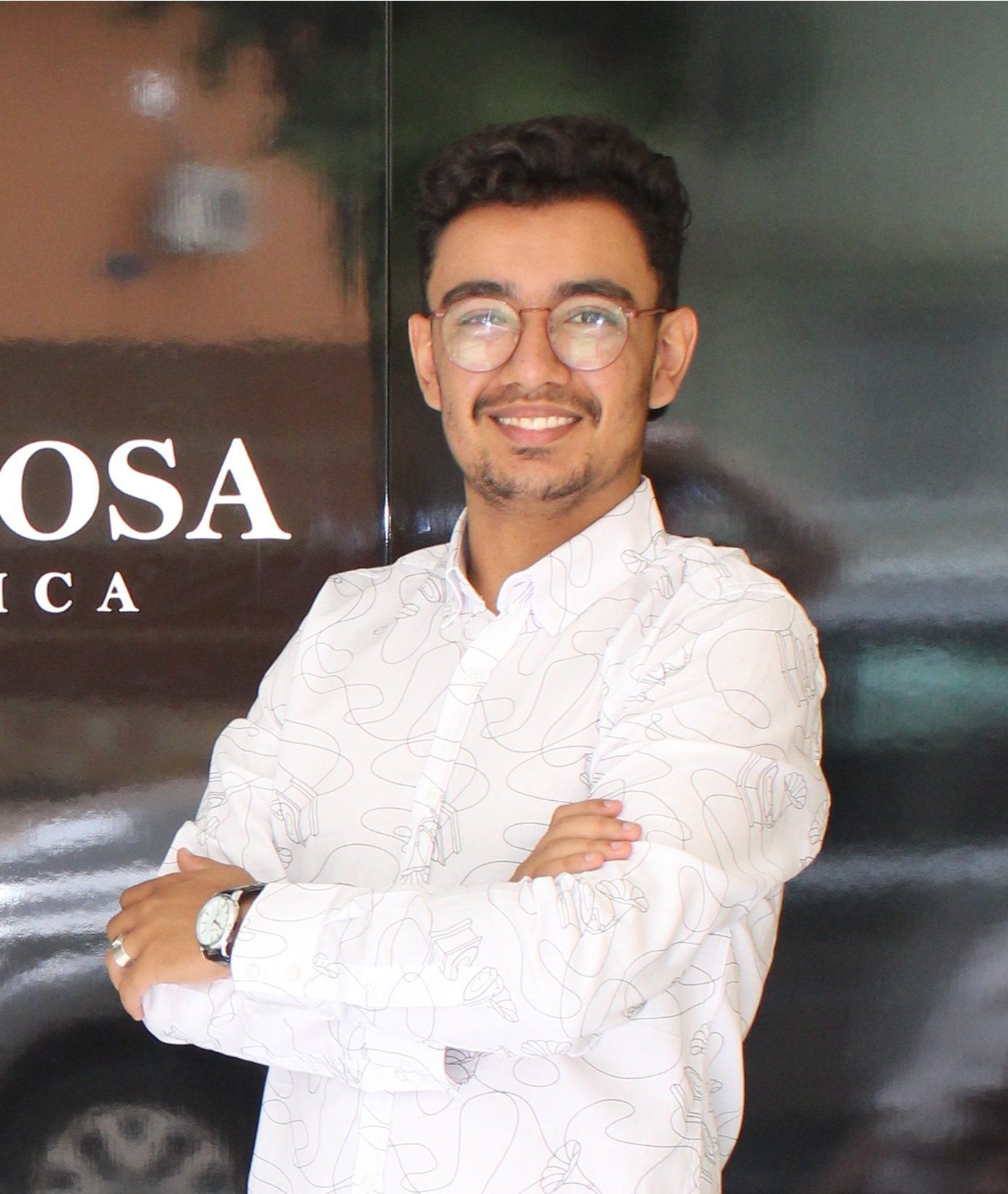 Um jovem de óculos e camisa branca está de pé com os braços cruzados.