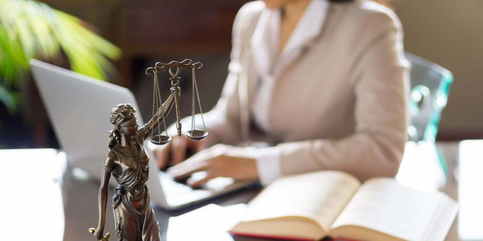 Uma mulher está sentada em uma mesa com uma estátua da justiça e um laptop.