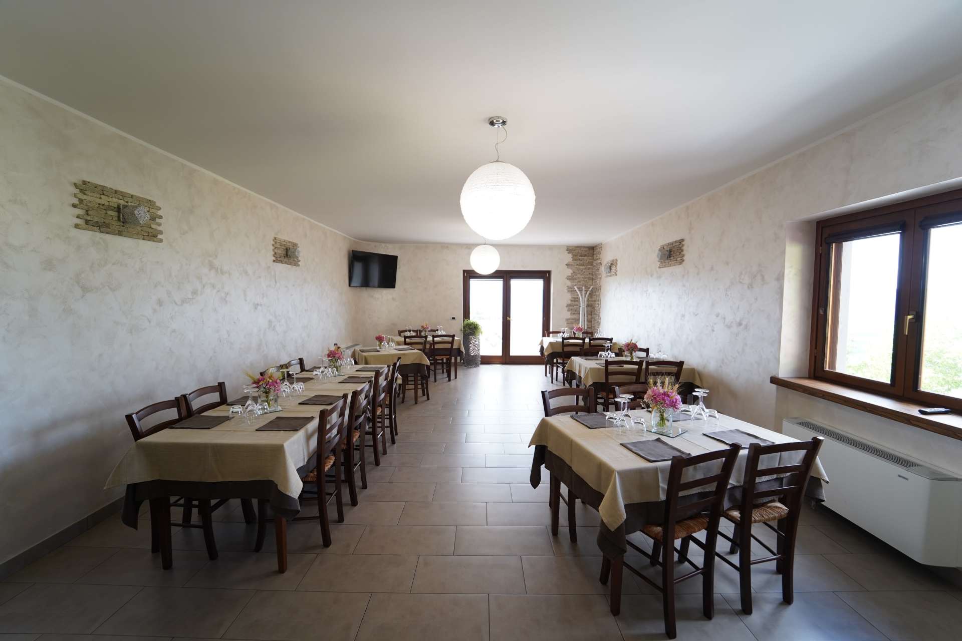 Sala da pranzo vuota con tavoli apparecchiati per un pasto, pareti bianche e grandi finestre.