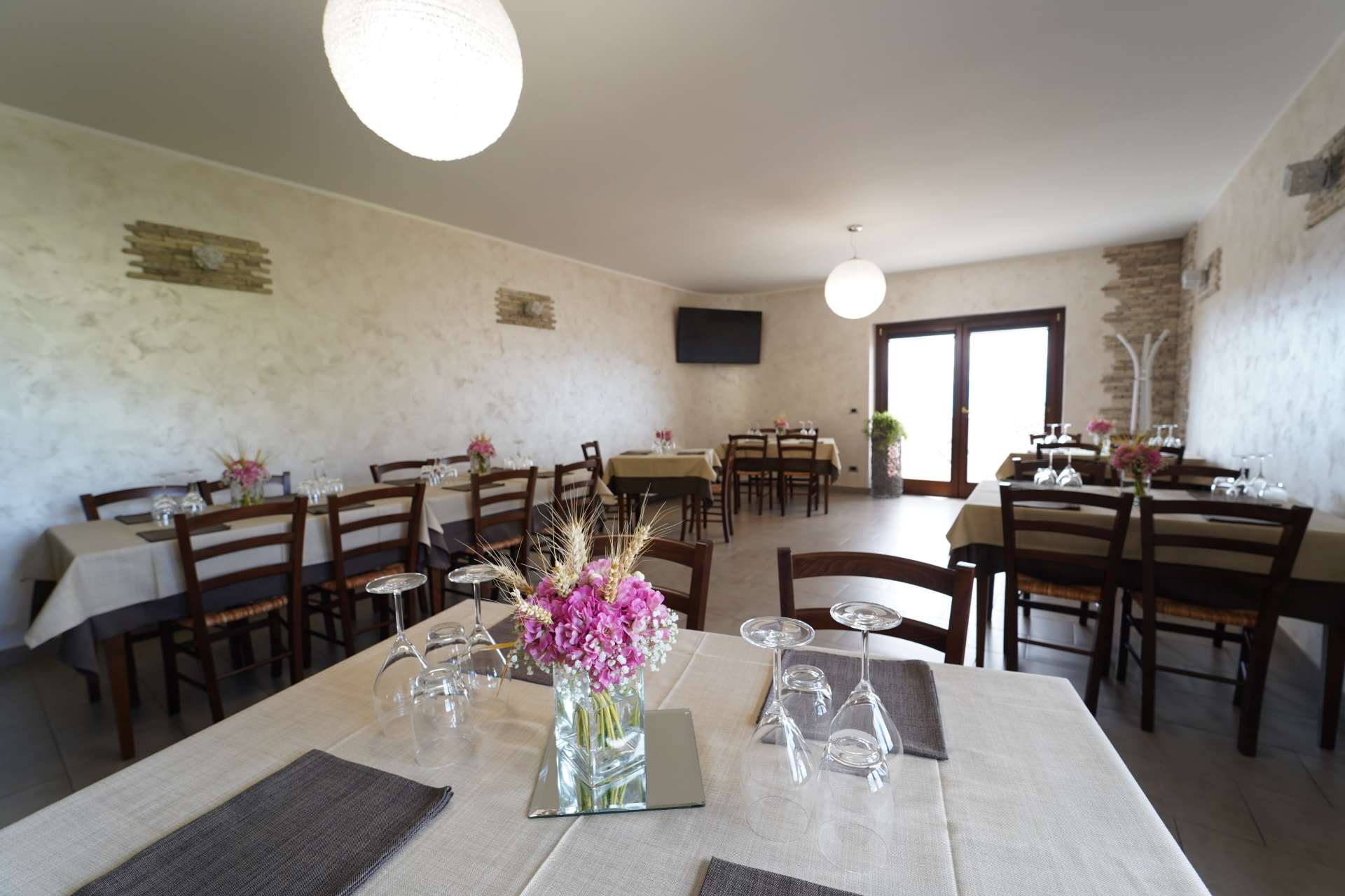 Luminosa sala da pranzo con tavoli bianchi, sedie in legno, lampade a sospensione e un centrotavola floreale vicino a una porta a vetri.