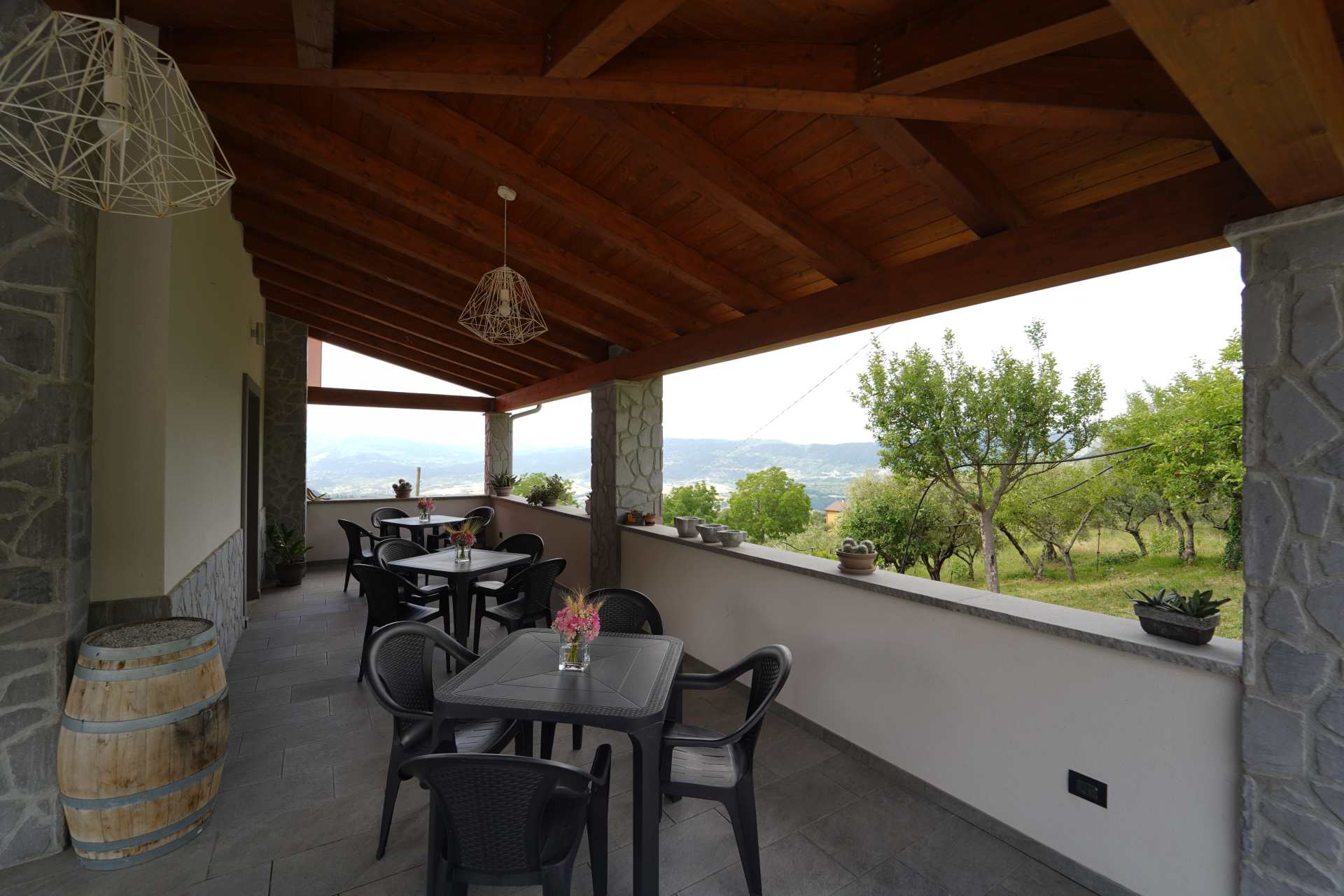 Patio coperto con tavoli e sedie neri, soffitto in legno e vista su alberi e colline.