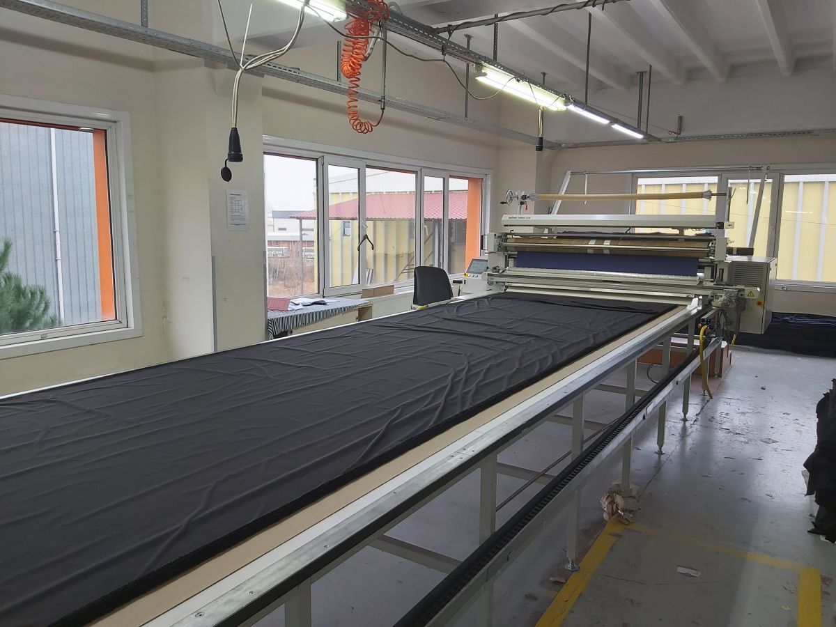 Grote industriële snijtafel in een werkplaats, met zwart textiel dat eronder onder felle plafondlampen ligt.