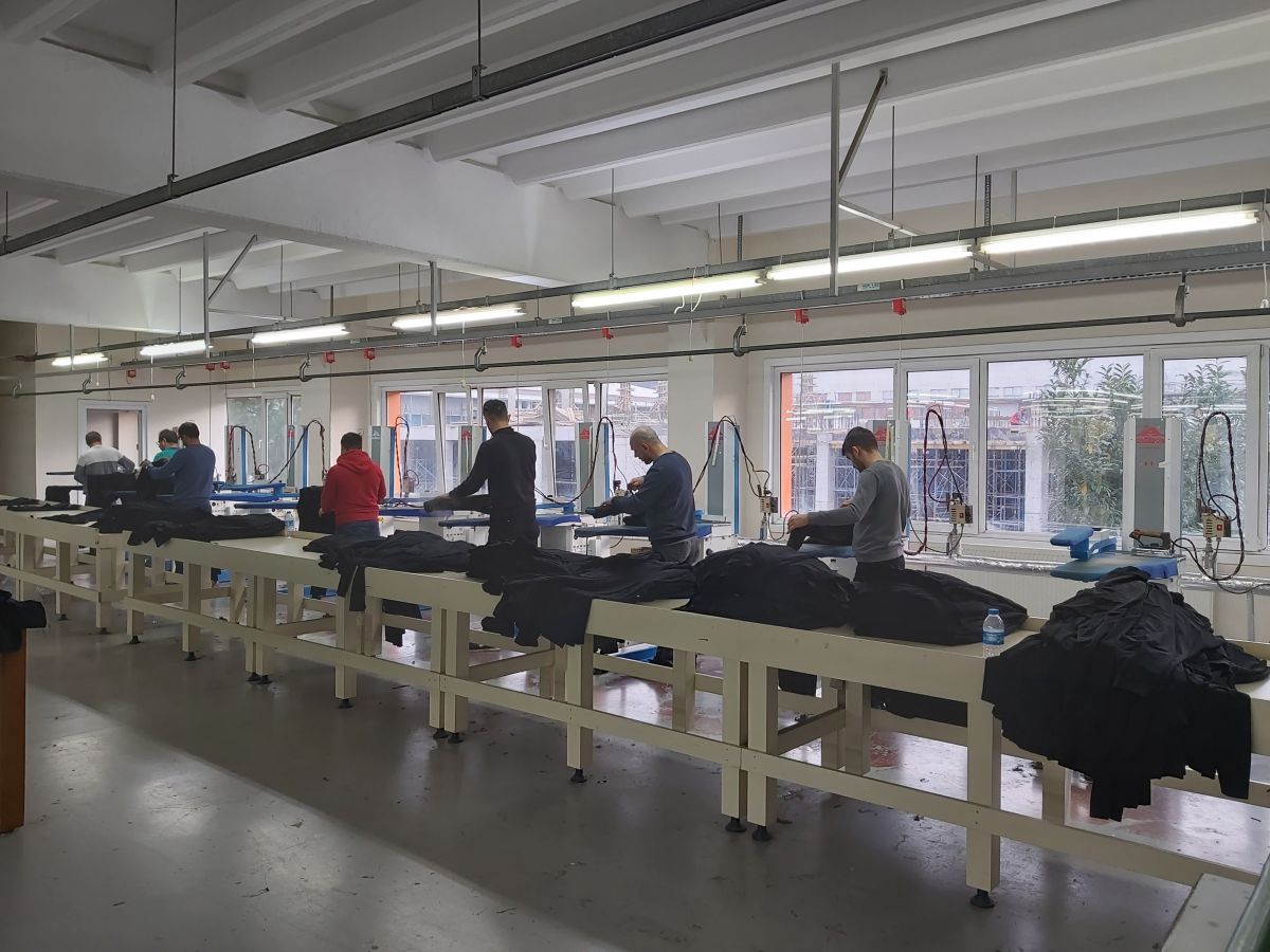 Mensen staan ​​aan lange werktafels in een lichte, industriële werkruimte met hanglampen en ramen.