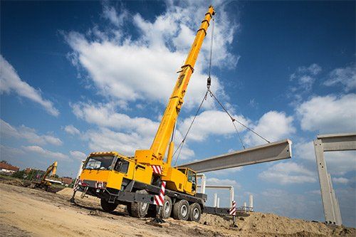 Yellow crane lifting a concrete beam at a construction site under a blue sky with clouds.