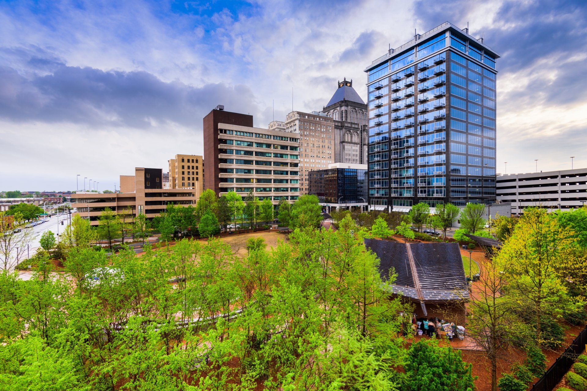 City skyline with modern high-rise buildings, green trees in the foreground, and overcast sky.