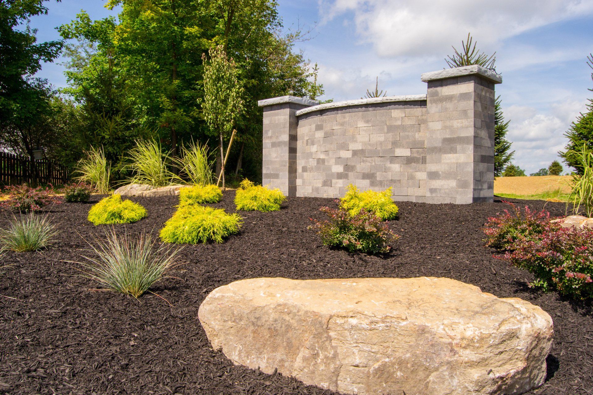 Landscape with a stone wall and various shrubs, set in black mulch, with a large rock in the foreground.