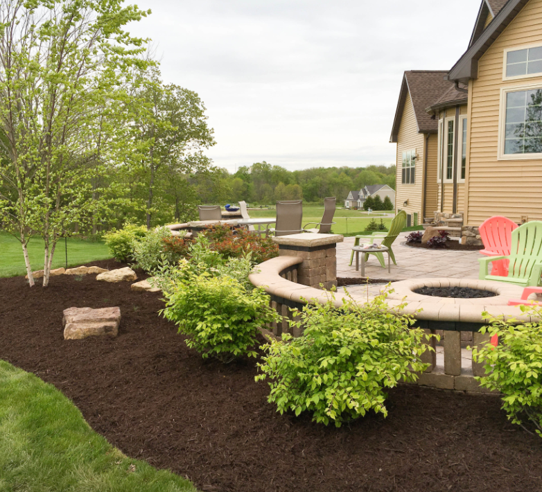 A backyard patio with a fire pit, seating, and landscaping next to a house under a cloudy sky.