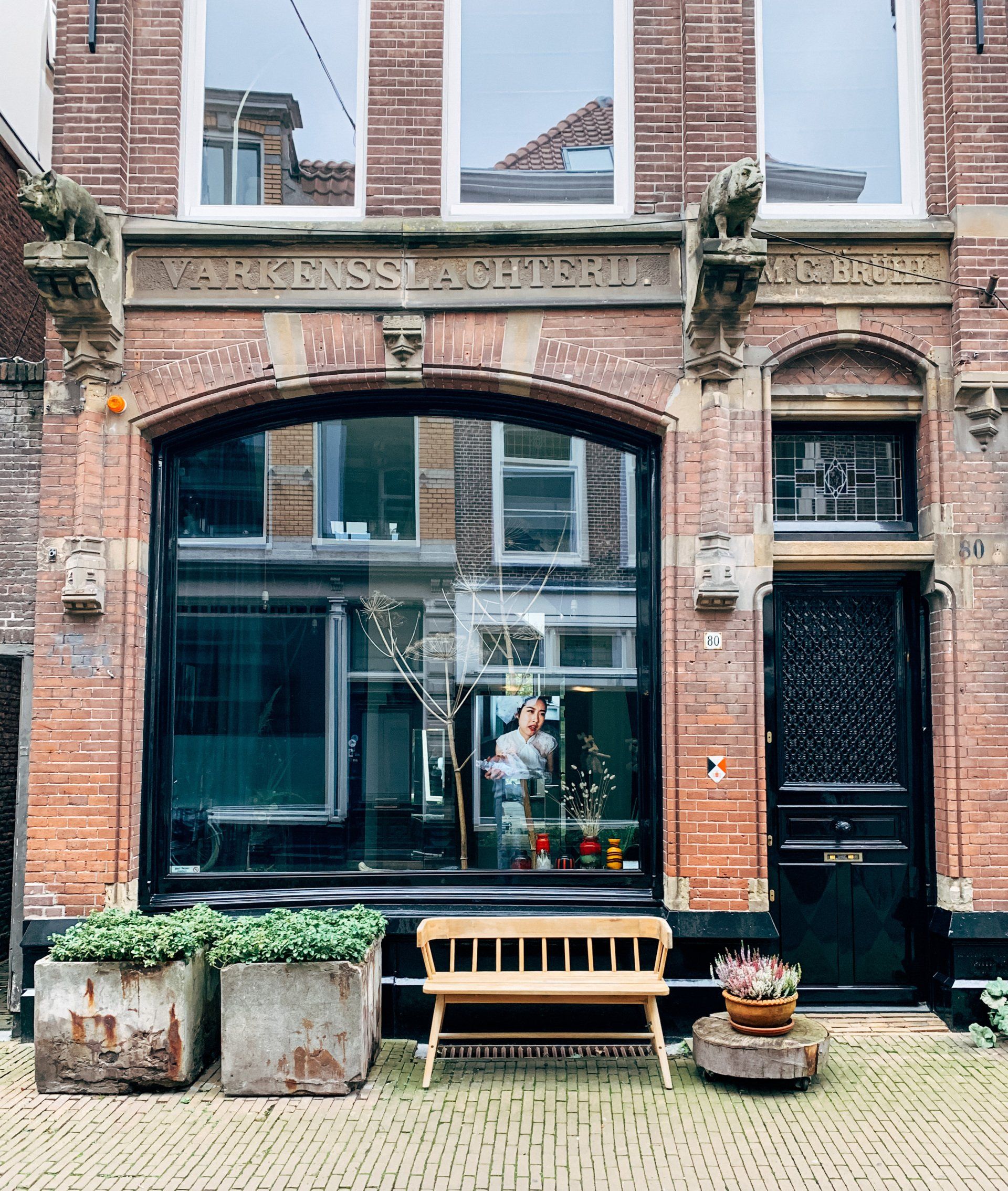 Gevel van een gebouw van rode baksteen met een groot raam, een zwarte deur, een bankje en plantenbakken.