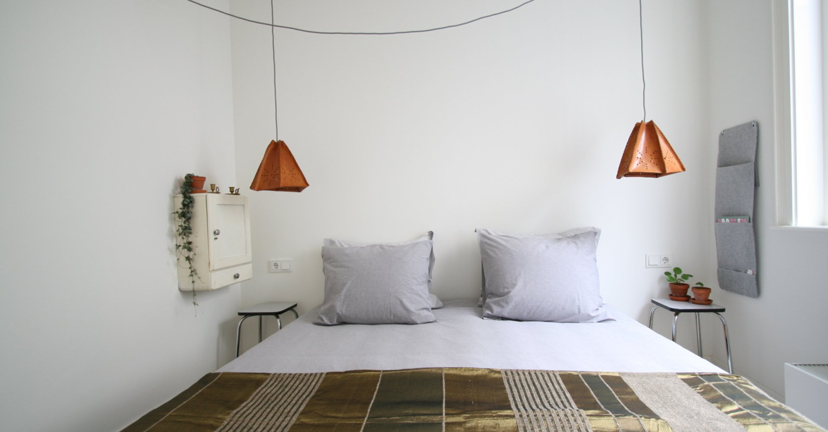 Slaapkamer met witte muren, twee koperkleurige hanglampen, een bed met grijze kussens en een deken met patroon.