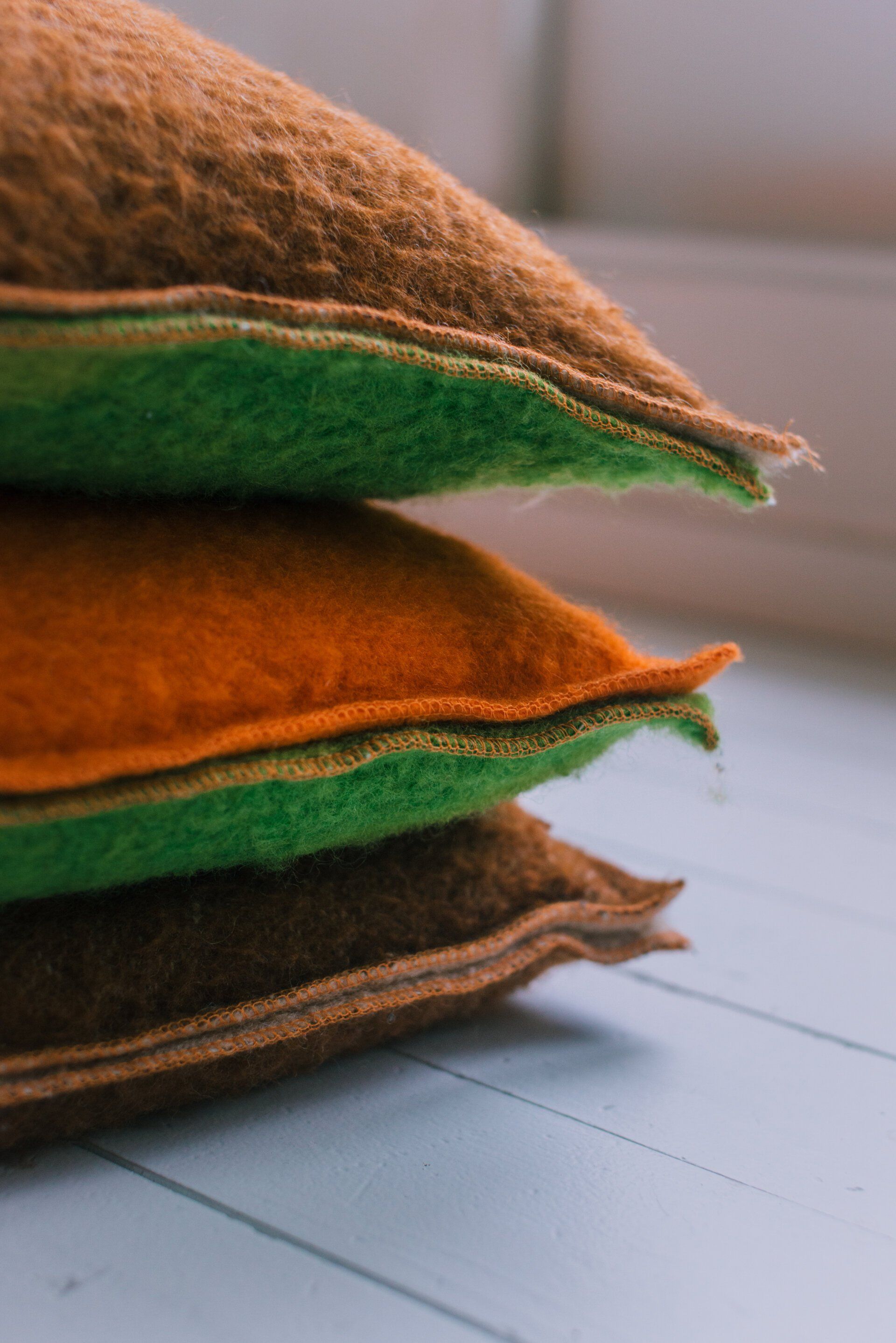 Een stapel vilten kussens: bruin, oranje en groen, die tegen een witte ondergrond leunen.