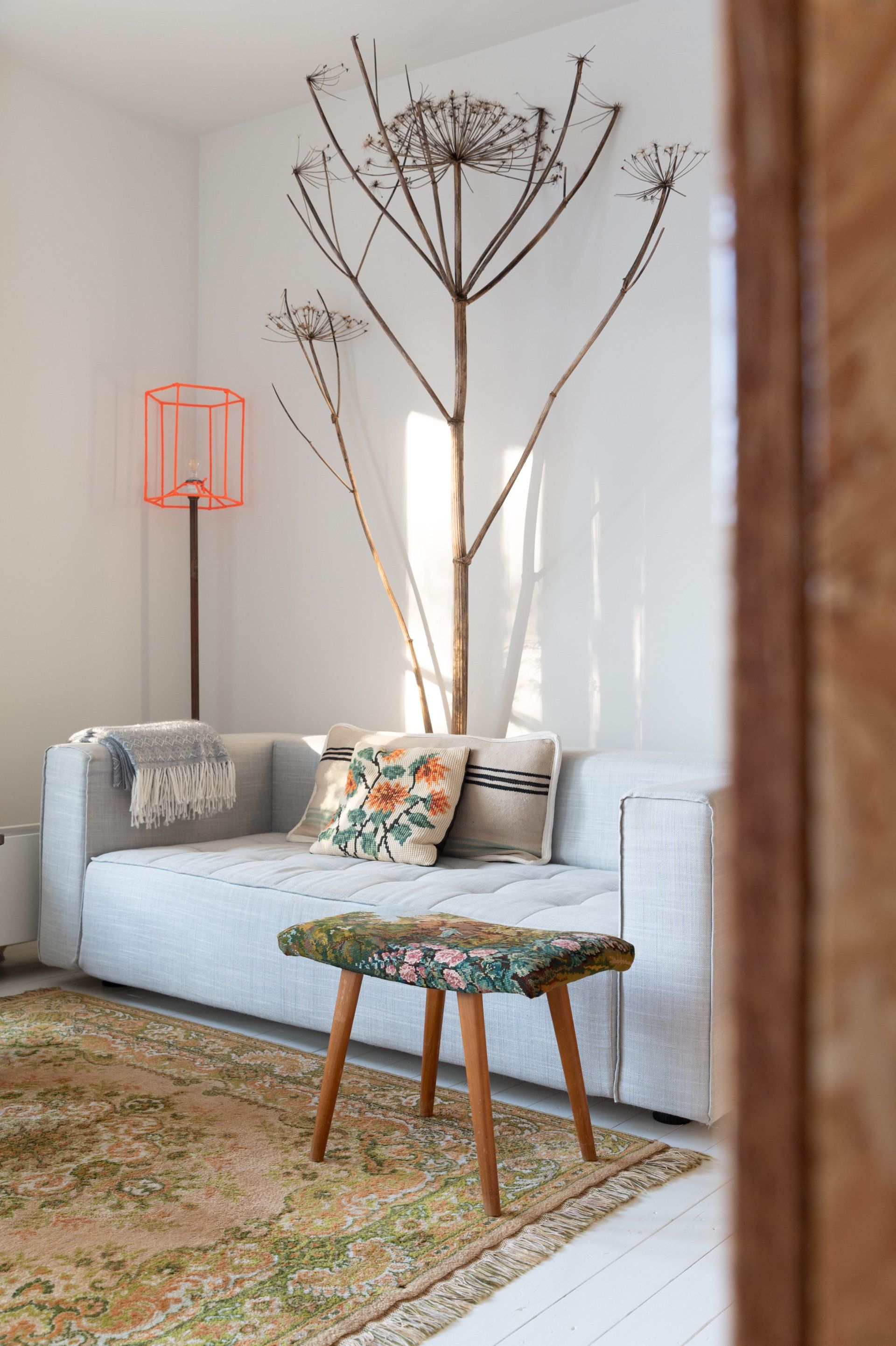 Een woonkamer met een grijze bank, een bloemenkleed en een krukje. Achter de bank staat een hoge, verdroogde plant.