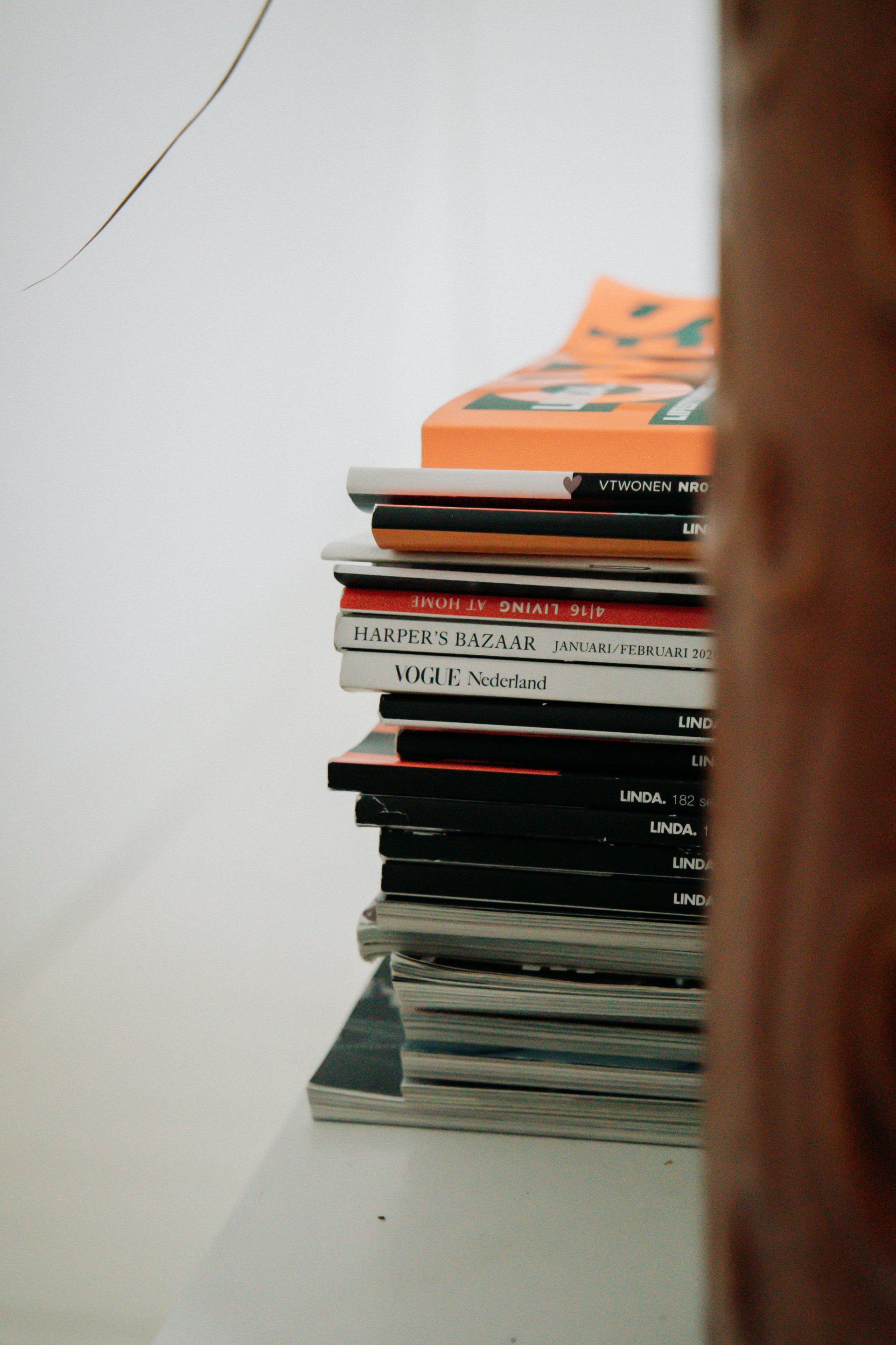 Een stapel boeken en tijdschriften op een witte plank. Bovenaan ligt een oranje boek.