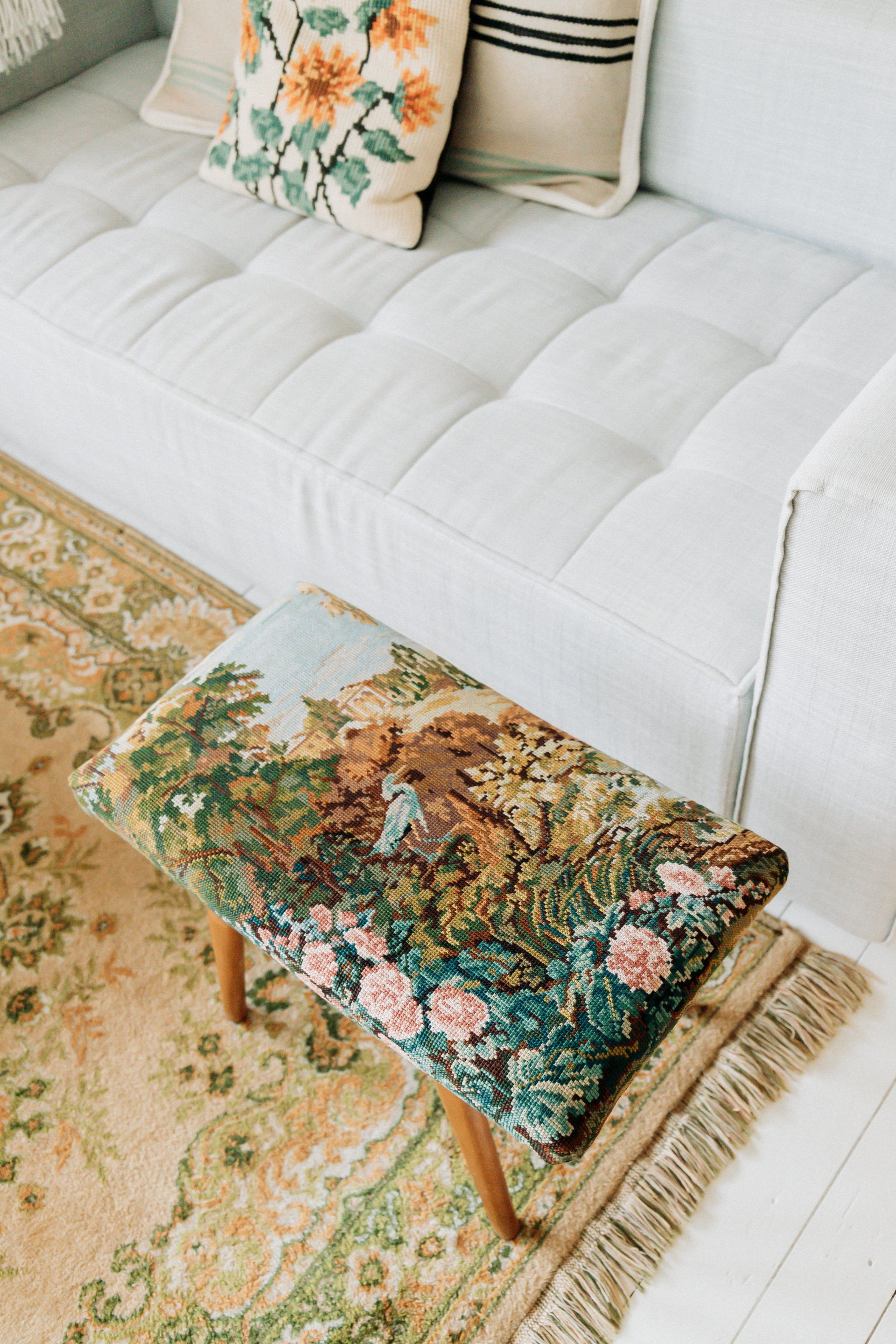 Een lichtgekleurde sofa met geborduurde bloemenkussens, een met wandtapijt beklede voetenbank en een bloemenkleed.